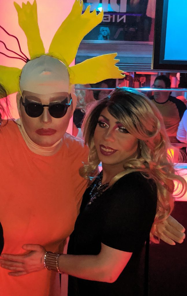 Image: Miss Kitsch (left) and DeCevia Mann (Right) posing together after a performance in Peoria IL. The neon of the night club glows behind them. Photo by the author.