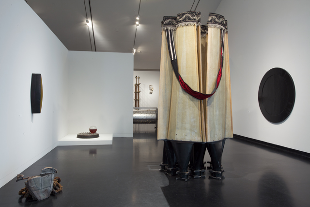 Image: Terry Adkins Recital, installation view, Tang Museum, 2012. The image shows several of Adkins' sculptural works. A large one and a much smaller one are free-standing on the floor of the gallery, two are mounted on the walls at either side of those sculptures, and several can be seen in the background, on pedestals and in other sections of the exhibition space. Photo courtesy of the Mary and Leigh Block Museum, Northwestern University.
