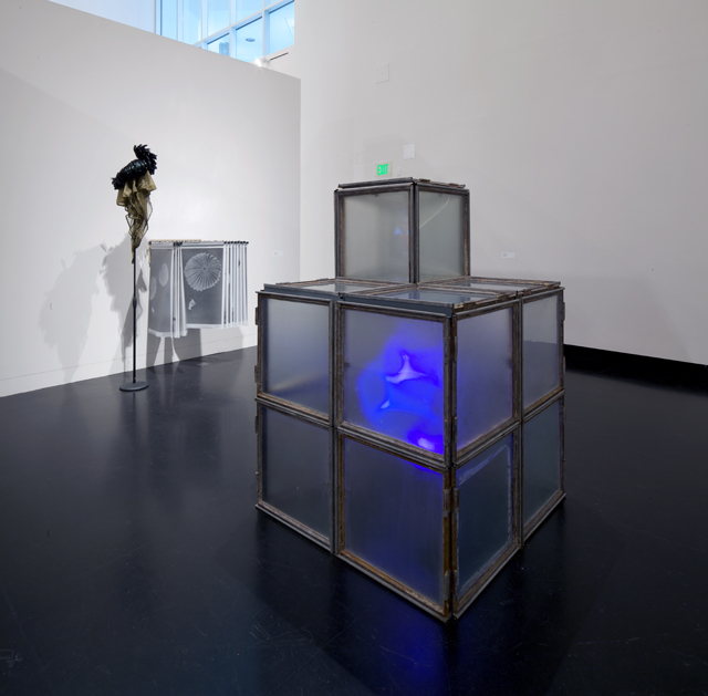 Image: Terry Adkins Recital, installation view, Tang Museum, 2012. One large, modular, square sculpture with translucent sides sits directly on the floor with a glowing blue light shining from within. In the background two sculptures can be seen, one on a stand and another mounted directly on the wall. Photo courtesy of the Mary and Leigh Block Museum, Northwestern University.