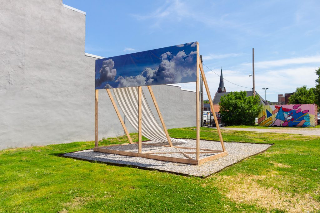 Image: Kahlil Robert Irving, MOBILE STRUCTURE; RELIEF & Memorial: (Monument Prototype for a Mass); 2019. Sculptural Installation. The structure is made up of wooden supports and with a banner of clouds going across the top, and a black and white striped banner going down the backside. Photo by Shabez Jamal. 