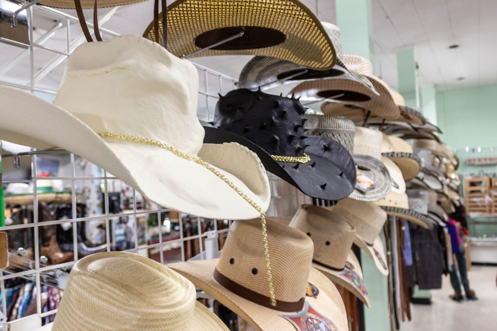 Image: Yowshien Kuo at Carrillo Western Wear, 2019. Installation with hand-painted apparel. A rack in a clothing store displays rows of cowboy hats. The hat in the middle is black with spikes. Photo by Shabez Jamal. 