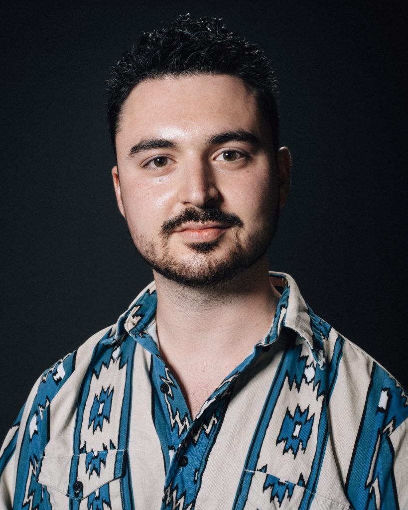 Image: A photograph of Jack Radley. He is looking forward at the viewer and wearing a shirt with a turquoise and tan southwest pattern.