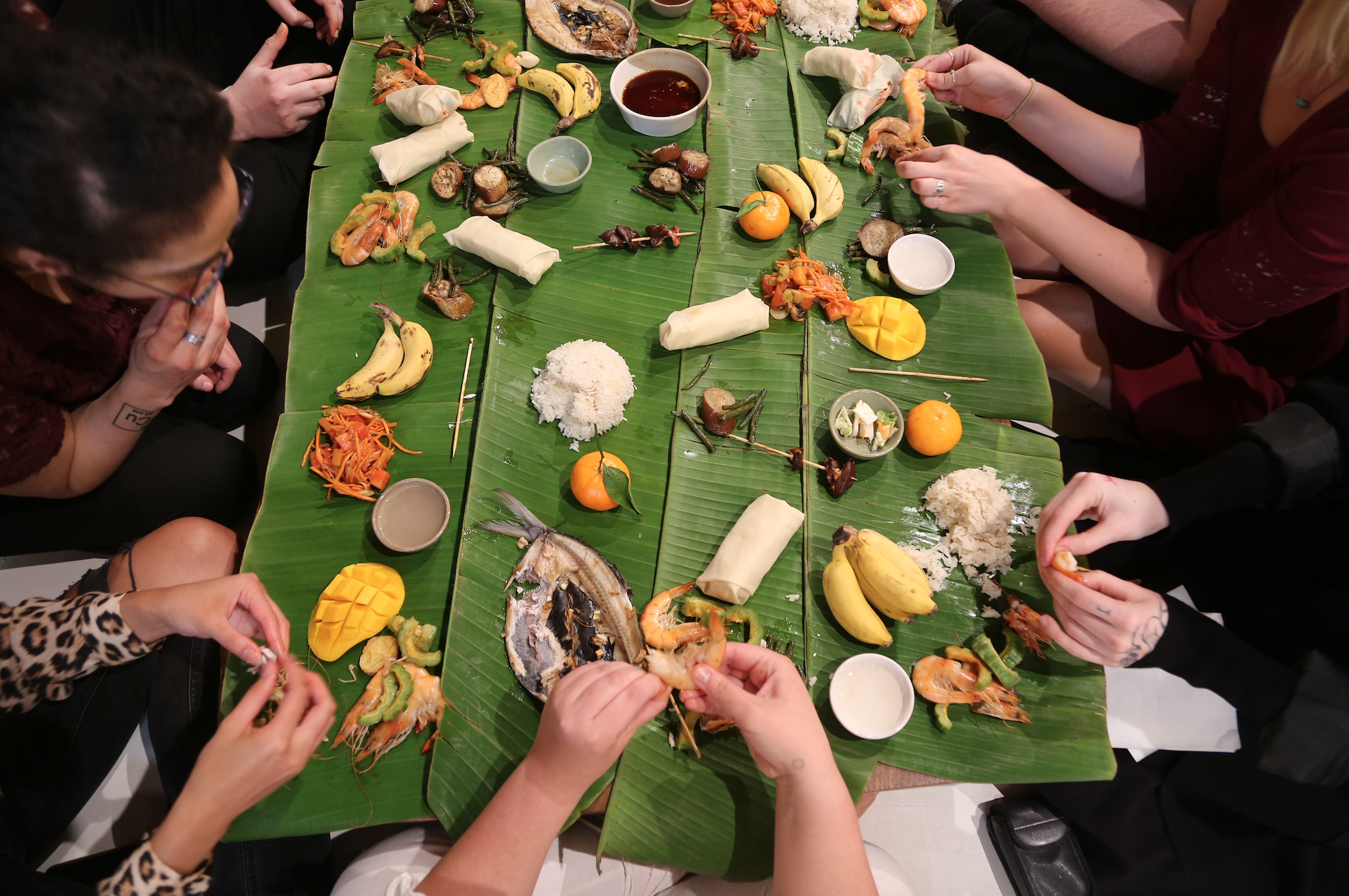 Image: Photo of Devyn's feast held at Ground Level Platform. The photo is taken from overhead and shows bright green banana leaves serving as a long table. On the leaves are various mounds and pieces of food such as a clump of white rice, halved and diced yellow mangoes, tangerines with a single green leaf still attached to them, and pairs of small and plump bananas in their peels. There are people all around the edges of the table touching the food with their hands, picking things apart. Photo courtesy of the artist.