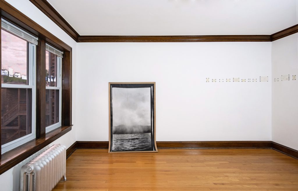 Image: Andre Keichian, 'Salt in the I' (installation view), 2019. A large black and white portrait photograph of a landscape mounted on wood curves against the base of the wall meeting the floor. To the left, a series of negatives of Keichian's family photographs adhered to the glass window. To the right, a series of the back, written sides of photographs placed across the corner of the room. Photo by Kim Becker. Image courtesy of Kyle Bellucci Johanson.