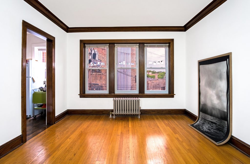 Image: Andre Keichian, 'Salt in the I' (installation view), 2019. A series of negatives of Keichian's family photographs adhered to the glass window. To the left, the entrance to Bellucci Johanson's kitchen. To the right, A large black and white portrait photograph of a landscape framed in oak wood curves against the base of the wall meeting the floor. Photo by Kim Becker. Image courtesy of Kyle Bellucci Johanson.