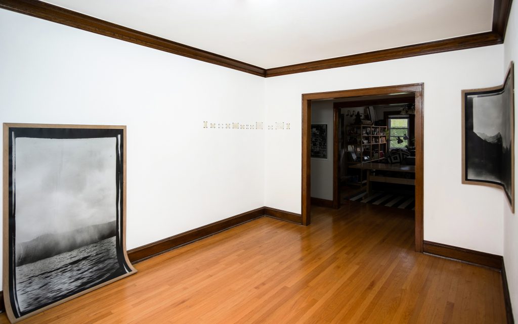 Image: Andre Keichian, 'Salt in the I' (installation view), 2019. A large black and white portrait photograph of a landscape mounted on wood curves against the base of the wall meeting the floor. To the right, a series of the back, written sides of photographs placed across the corner of the room; a large black and white landscape photograph mounted on wood curves against the corner of the room, as part of the exhibition installation at table. In the background, a view of Bellucci Johanson's living room through the entryway of the dining room. Photo by Kim Becker. Image courtesy of Kyle Bellucci Johanson.