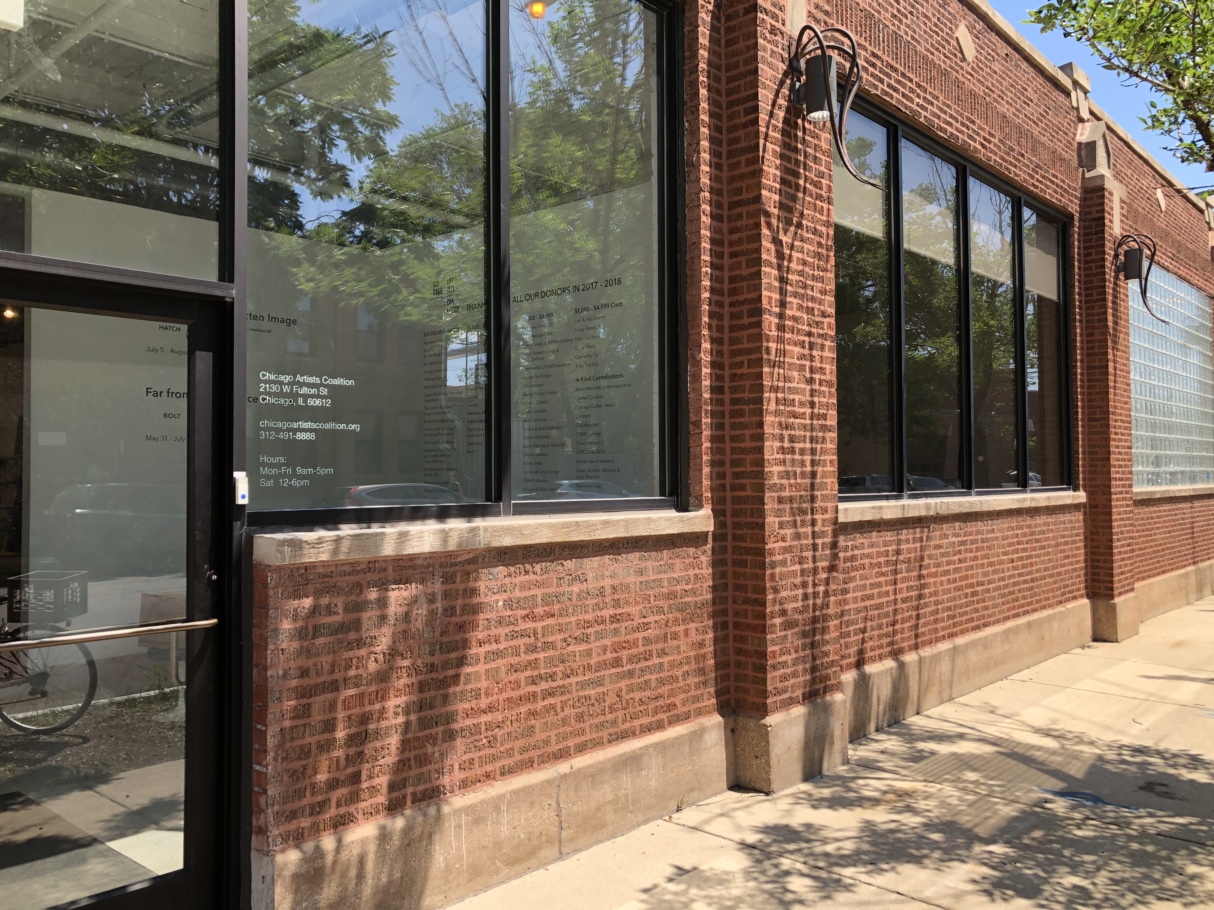 Image: An external shot of Chicago Artists Coalition. The building is made up of red bricks and large, rectangular windows. The entryway of the gallery can be seen through the front door. The sun is shining and the windows reflect the blue sky and leafy green trees that line the sidewalk. Photo courtesy of Chicago Artists Coalition.