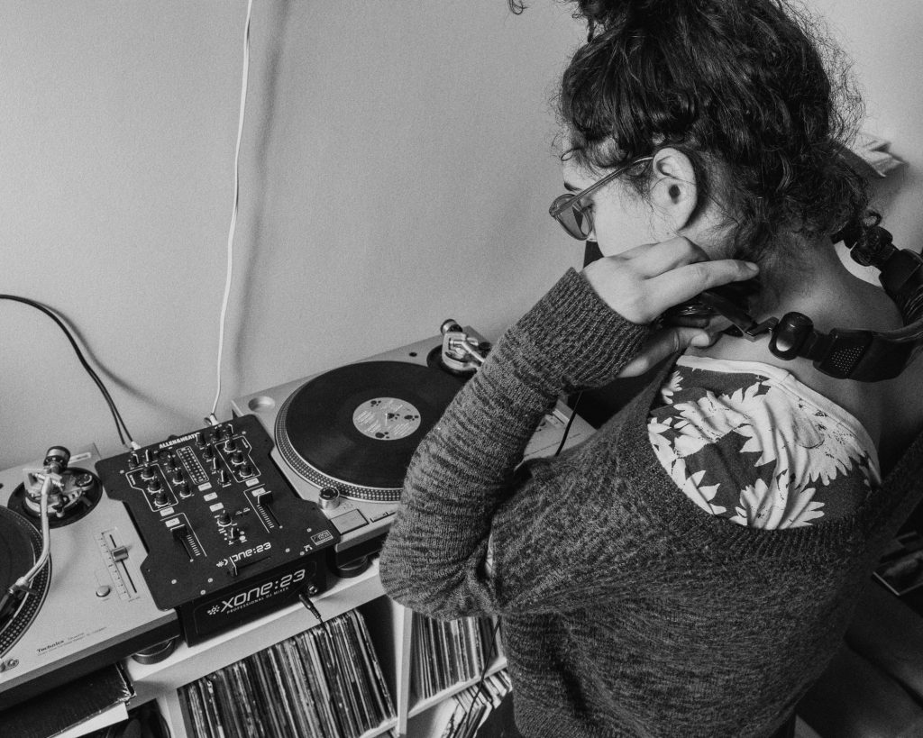Image: In a black-and-white photo, Ariel looks down at the turntables and mixer that sit on a shelf full of records in her living room. Photo by Ryan Edmund.