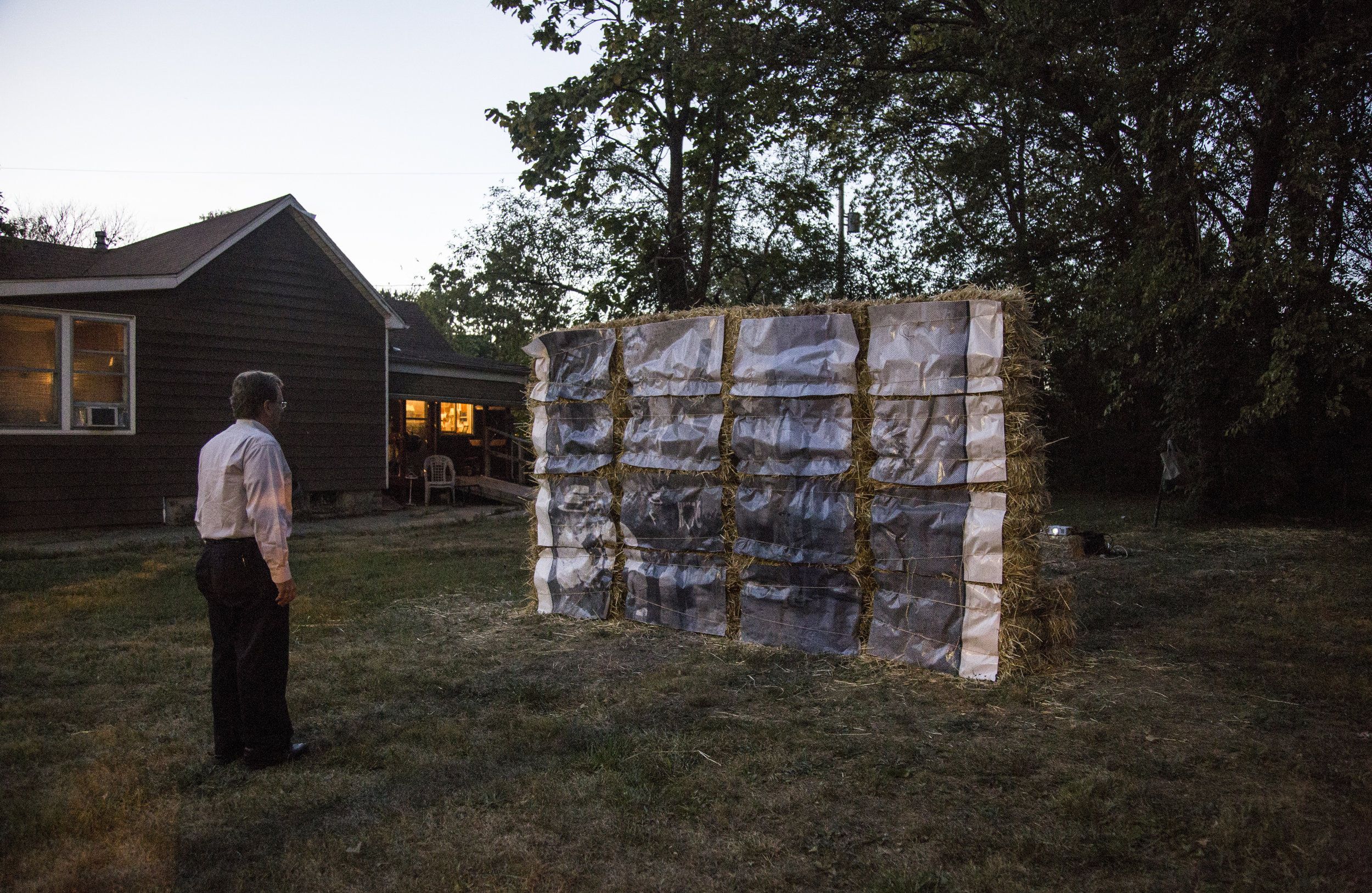 Image: Documentation image of Cass Davis’ Witness, 2017 taken at sunset. The piece is made up of 30 hay bales, bailing twine, and an archival reproduction of a photograph from the 1908 Springfield Race Riot. Hay bales are stacked horizontally and vertically to achieve height. The archival photograph has been enlarged and cut into 16 even-sized rectangles and attached to the hay bales with twine. The piece is displayed on a vacant lot of grass with trees in the background and a house in the neighboring lot. A man wearing black pants and a long sleeve collared shirt faces the installation. Photo by the artist. Archival photograph courtesy of the University of Illinois, Springfield Archives.