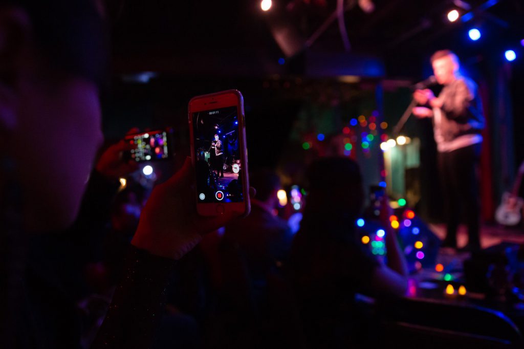 Image: Chelsea Fiddyment performing at Dan’s Silverleaf in Denton, Texas. Fiddyment appears fuzzy in the background, in full profile, standing on stage and speaking into a microphone while looking out at the audience. White stage lights and colorful stringed lights are above and below Fiddyment. The camera looks up at Fiddyment from the vantage-point of the audience and, in the foreground, audience members hold up their phones to video-record Fiddyment. Fiddyment wears a dark-colored bomber jacket, a light shirt, and dark pants. Photo by Leah Jones. Courtesy of the artist.