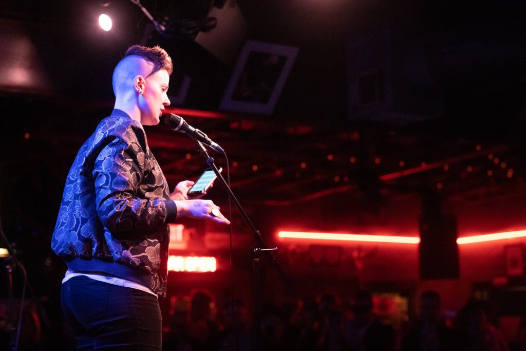  Image: Chelsea Fiddyment performing at Spiderweb Salon’s New Year’s Revolution show, at Dan’s Silverleaf in Denton, Texas, on January 12, 2019. Fiddyment appears in profile, standing on stage and speaking into a microphone while looking out at the audience. The camera looks up at Fiddyment from a low angle, such that part of the ceiling is visible, including a white stage light on the left side of the frame and red lights above the audience area on the right side of the frame. Fiddyment wears a dark-colored, floral-print bomber jacket and dark pants. Photo by Nina Chantanapumma. Courtesy of the artist.