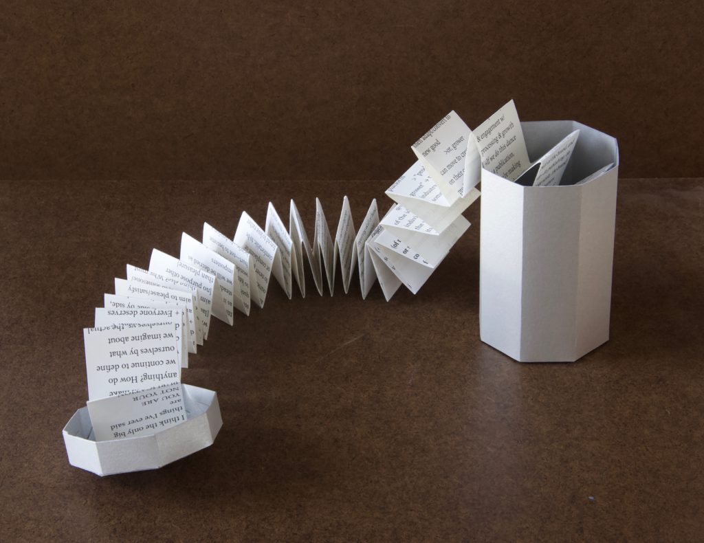 Image: A photo of “Trash Baby Manifesto,” a book by Chelsea Fiddyment, 2018. The book’s casing looks like a trashcan — octagonal at the base and top, with a lid. It is made out of thin white cardboard. The lid and can sit on opposite sides of the frame. In this image, the book’s contents — a length of accordion-folded paper, long and narrow, attached at one end to the lid and continuing into the “trashcan” at the other end — stretches across the dark surface between them. The paper is white with black serif text in a small font. Photo by Meekling Press. Courtesy of the artist.
