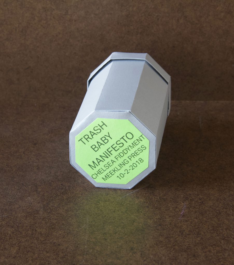 Image: A photo of “Trash Baby Manifesto,” a book by Chelsea Fiddyment, 2018. The book’s casing looks like a trashcan — octagonal at the base and top, with a lid. It is made out of thin white cardboard. The focal point of the image is the bottom of the trashcan, where a light green octagonal sticker reads, in all capital letters, “Trash Baby Manifesto, Chelsea Fiddyment, Meekling Press, 10-2-2018.” The book sits on a dark surface. Photo by Meekling Press. Courtesy of the artist.