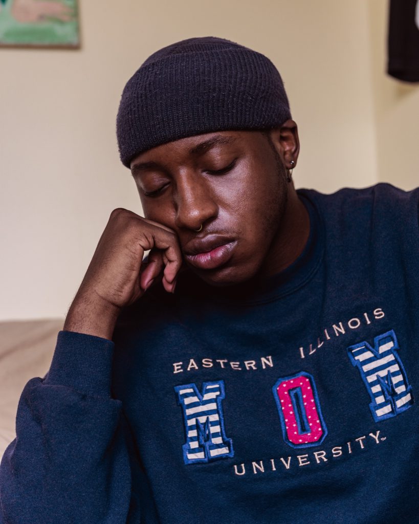 Image:Miss Twink USA closes his eyes and rests his chin on his fist. He's wearing a beanie and a navy blue sweatshirt that reads, "Eastern Illinois University Mom." Photo by Ryan Edmund.