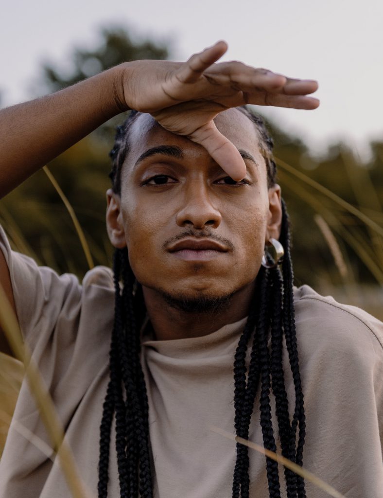 Image: Sitting in tall grasses, Morenxxx stares directly at the camera, with their hand partially covering part of their face. Photo by Ryan Edmund.