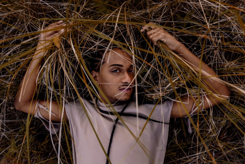 Image: Morenxxx lays in tall grasses and stares directly into the camera. Photo by Ryan Edmund.