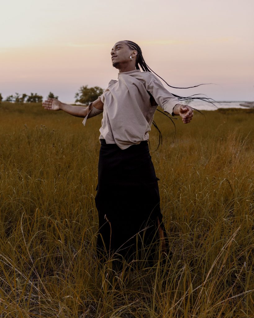 Image: Morenxxx stands in the grasses with their arms outstretched and whips their braids around their head. Photo by Ryan Edmund.