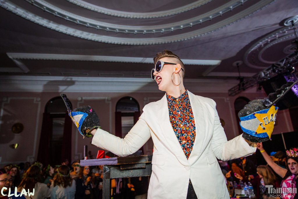 Image: Chelsea Fiddyment as Sir Elton Brawn, celebrating a championship win at CLLAW XXXII (Chicago League of Lady Arm Wrestlers), at Logan Square Auditorium in 2019. The camera looks up at Fiddyment from the vantage-point of the audience. Fiddyment is well-lit in the middle of a large, dim room that has high windows and a white ceiling with ornamental mouldings. Behind Fiddyment, dozens of the audience members are partially visible, most cheering and wearing sparkly headwear. On each hand, Fiddyment wears a large foam fist, with the middle finger raised, and Fiddyment stands with arms out, as if giving the middle finger in celebration to both sides of the room. Fiddyment’s mouth is open as if yelling, and an artificial, painted gap is visible between Fiddyment’s two front teeth. Fiddyment wears sparkly sunglasses, a light-colored sportcoat, and a bright, floral-print shirt buttoned to the top. The image has logos for “CLLAW” and “Trainman Photography” in its bottom corners. Photo by Trainman Photography. Courtesy of the artist.