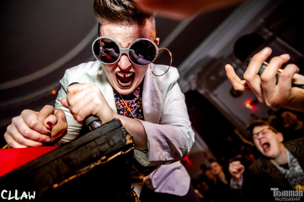 Image: Chelsea Fiddyment as Sir Elton Brawn, winning an arm-wrestling match at CLLAW XXXII (Chicago League of Lady Arm Wrestlers), at Logan Square Auditorium in 2019. In this close-up, the camera looks up at Fiddyment. Fiddyment is well-lit near the center of the frame, in the midst of, and seemingly about to win, an arm-wrestling match. Fiddyment’s other hand grips a handle mounted to the table. Fiddyment’s mouth is open as if yelling, and an artificial, painted gap is visible between Fiddyment’s two front teeth. Fiddyment wears sparkly sunglasses, a light-colored sportcoat, and a bright, floral-print shirt buttoned to the top. The image has logos for “CLLAW” and “Trainman Photography” in its bottom corners. Photo by Trainman Photography. Courtesy of the artist.