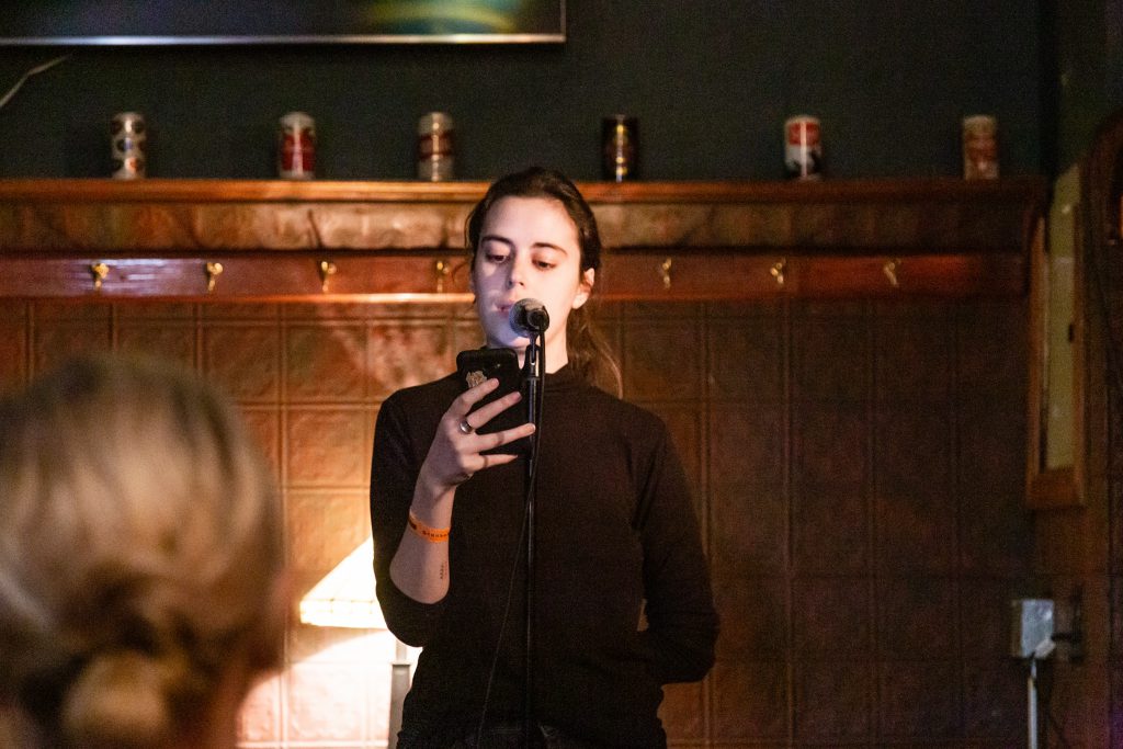 Image: Molly Gunther performing at Unreal at Schubas. Gunther stands at the front of the room, speaking into a microphone while looking past it at a phone. Behind Gunther is a copper-colored wall, made of a grid of low-relief tiles; above that are several decorative beer cans on a ledge and a dark green section of wall. Gunther wears a black with a sleeve rolled up. The back of an audience member’s head is unfocused in the foreground. Photo by Joshua Clay Johnson.