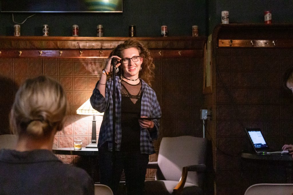 Image: Terry Galvan performing at Unreal at Schubas. Galvan stands at the front of the room, smiling out at the audience, with one hand around the microphone. Behind Galvan is a copper-colored wall, made of a grid of low-relief tiles, and a door; above that are several decorative beer cans on a ledge and a dark green section of wall. Chelsea Fiddyment is partially visible in the background, cueing up a song on a laptop. Galvan wears a blue plaid shirt open over a lacy black shirt, dark pants, and dark-rimmed glasses. The back of an audience member’s head is unfocused in the foreground. Photo by Joshua Clay Johnson.