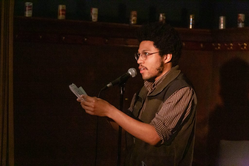 Image: Crum the Villain performing at Unreal at Schubas. In this oblique, medium-length shot, Crum stands at the front of the room, small notebook in hand, speaking into a microphone while looking out at the audience. Behind Crum is a copper-colored wall, made of a grid of low-relief tiles; above that are several decorative beer cans on a ledge and a dark green section of wall. Crum wears a brown and white striped collared shirt with the sleeves rolled up, with a dark green vest half-zipped over that, as well as round, silver-rimmed glasses. Photo by Joshua Clay Johnson.