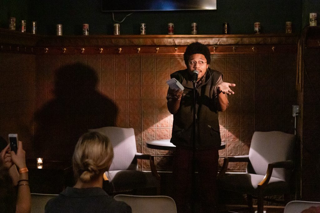 Image: Crum the Villain performing at Unreal at Schubas. Crum stands at the front of the room, notebook in hand, speaking into a microphone while looking at one side of the room. Crum’s arms are shrugged and eyebrows raised. Behind Crum is a copper-colored wall, made of a grid of low-relief tiles; above that are several decorative beer cans on a ledge and a dark green section of wall. Crum wears a brown and white striped collared shirt with the sleeves rolled up, with a dark green vest half-zipped over that. The backs of some audience members’ heads are fuzzy in the foreground. Photo by Joshua Clay Johnson.