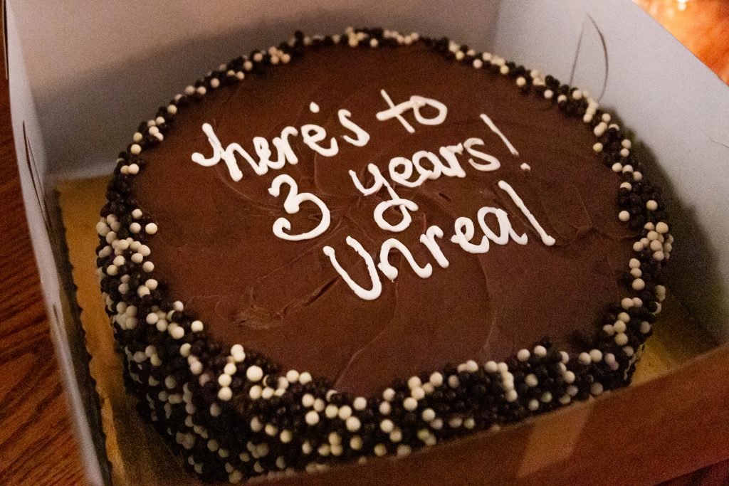  Image: A cake reading “here’s to 3 years! Unreal” in white icing. The cake is round and layered, with dark brown frosting and dark brown and white candies around its sides. It sits in an open box, a wooden table slightly visible beneath it. Photo by Joshua Clay Johnson.