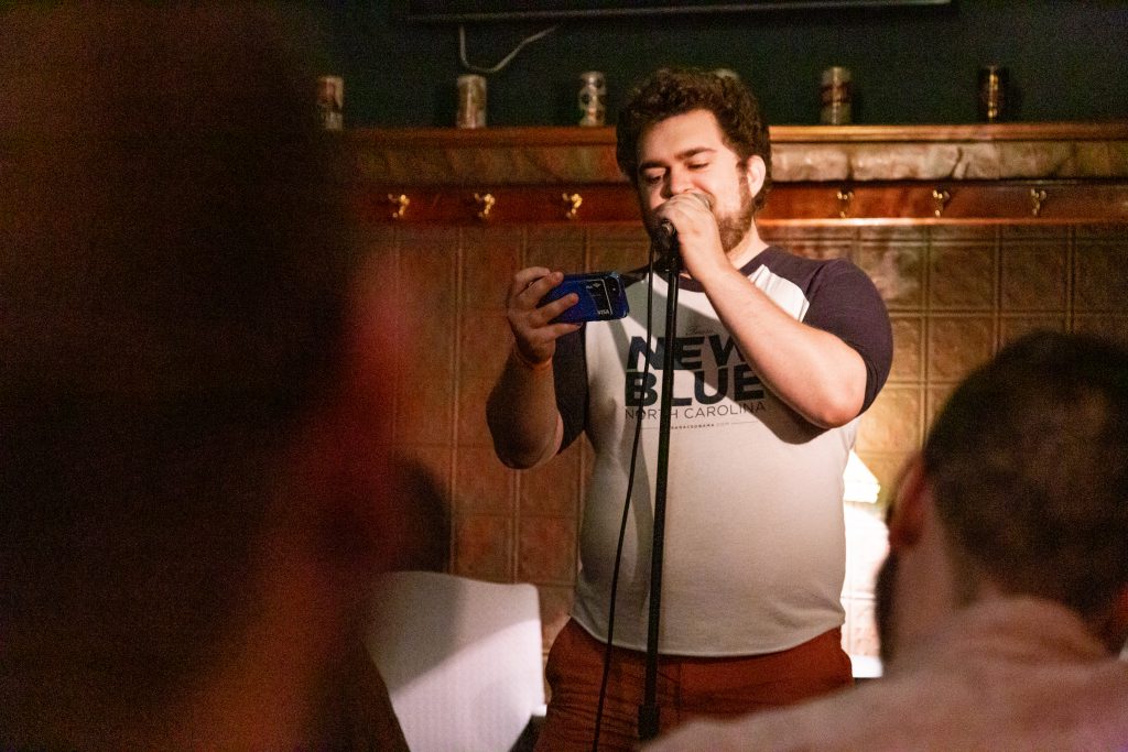 Image: Nils Robbins performing at Unreal at Schubas. Robbins stands at the front of the room, speaking into a microphone while reading off a phone. Behind Robbins is a copper-colored wall, made of a grid of low-relief tiles; above that are several decorative beer cans on a ledge and a dark green section of wall. Robbins wears burnt-orange pants and a navy and white baseball tee that reads “New Blue” and “North Carolina.” The backs of some audience members’ heads are unfocused in the foreground. Photo by Joshua Clay Johnson.