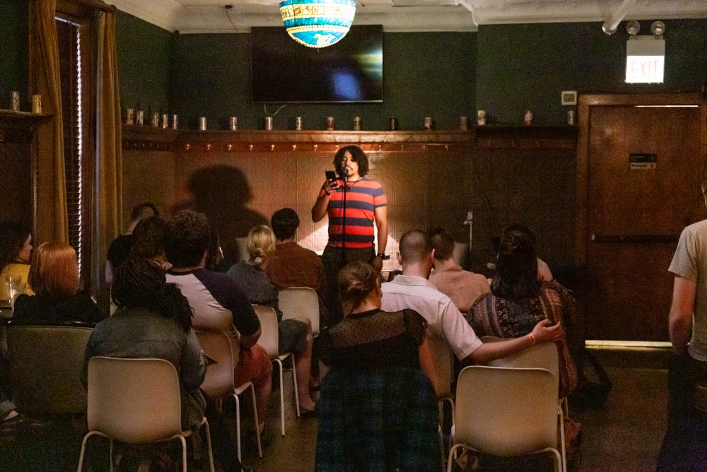 Image: Michael Tucker performing at Unreal at Schubas. In this long shot, showing half of the room, Tucker stands at the front, speaking into a microphone while reading off a phone. Behind Tucker is a copper-colored wall, made of a grid of low-relief tiles; above that are several decorative beer cans on a ledge, a dark green section of wall, and a flat-screen monitor. A stained glass light hangs from the ceiling. Tucker wears a red and blue striped tee with jeans. The backs of several audience members and their chairs are in the foreground. Photo by Joshua Clay Johnson.