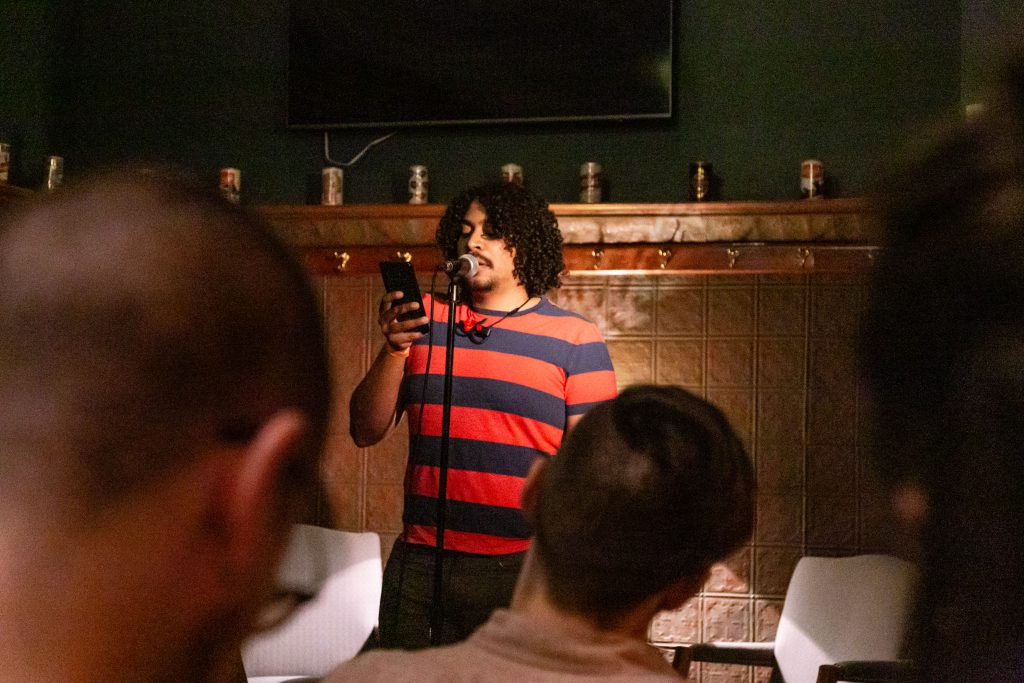 Image: Michael Tucker performing at Unreal at Schubas. Tucker stands at the front of the room, speaking into a microphone while reading off a phone. Behind Tucker is a copper-colored wall, made of a grid of low-relief tiles; above that are several decorative beer cans on a ledge, a dark green section of wall, and a flat-screen monitor. Tucker wears a red and blue striped tee with jeans. The backs of some audience members’ heads are unfocused in the foreground. Photo by Joshua Clay Johnson.