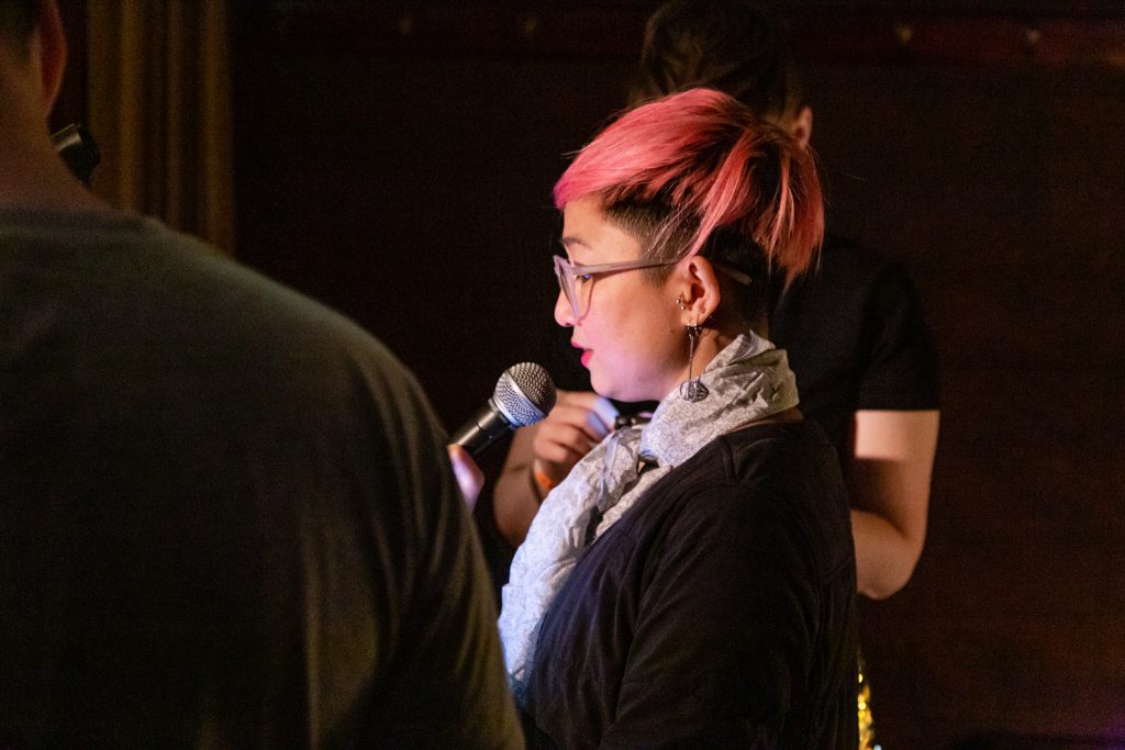 Image: Dao Nguyen performing at Unreal at Schubas. Nguyen stands in profile, lit brightly and speaking into the microphone. Two other performers, standing on either side of her, are only partially visible in the frame. Nguyen wears a black shirt with a silver scarf and pink glasses a few shades lighter than her hair. Photo by Joshua Clay Johnson.