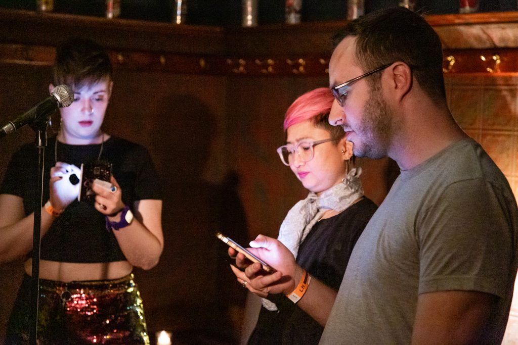 Image: Chelsea Fiddyment, Dao Nguyen, and John Wilmes performing a short play by Nguyen, at Unreal at Schubas. In this oblique, medium-length shot, all three performers stand at the front of the room, a microphone on a stand between them, each reading off or following along on their phone. Behind them is a copper-colored wall, made of a grid of low-relief tiles; above that are several decorative beer cans on a ledge and a dark green section of wall. Fiddyment wears a black cropped tee and red and gold sequined shorts; Nguyen wears a black shirt with a silver scarf and pink glasses a few shades lighter than her hair; and Wilmes wears a grey tee and dark-rimmed glasses. Photo by Joshua Clay Johnson.