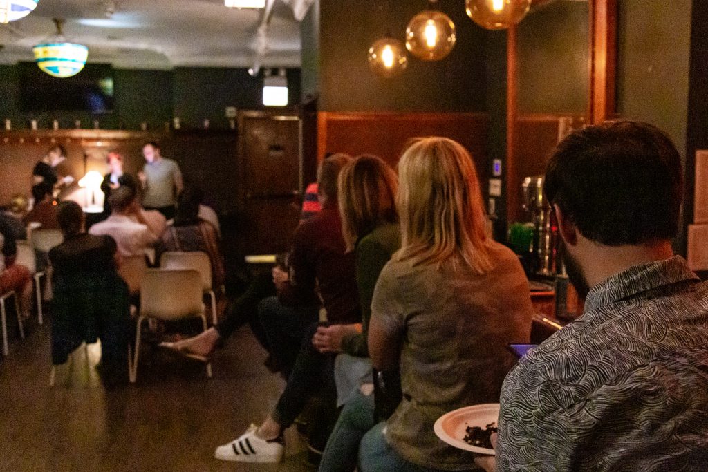 Image: The upstairs space at Schubas, showing Chelsea Fiddyment, Dao Nguyen, and John Wilmes performing at Unreal. The performers are fuzzy in the background, reading off their phones, while audience members in chairs watch. In the foreground, other audience members sit on stools at the bar, their backs to the camera. The walls are copper-colored at the bottom and dark green at the top. Stained glass lights hang above the main space and globular lights hang above the bar. Photo by Joshua Clay Johnson.