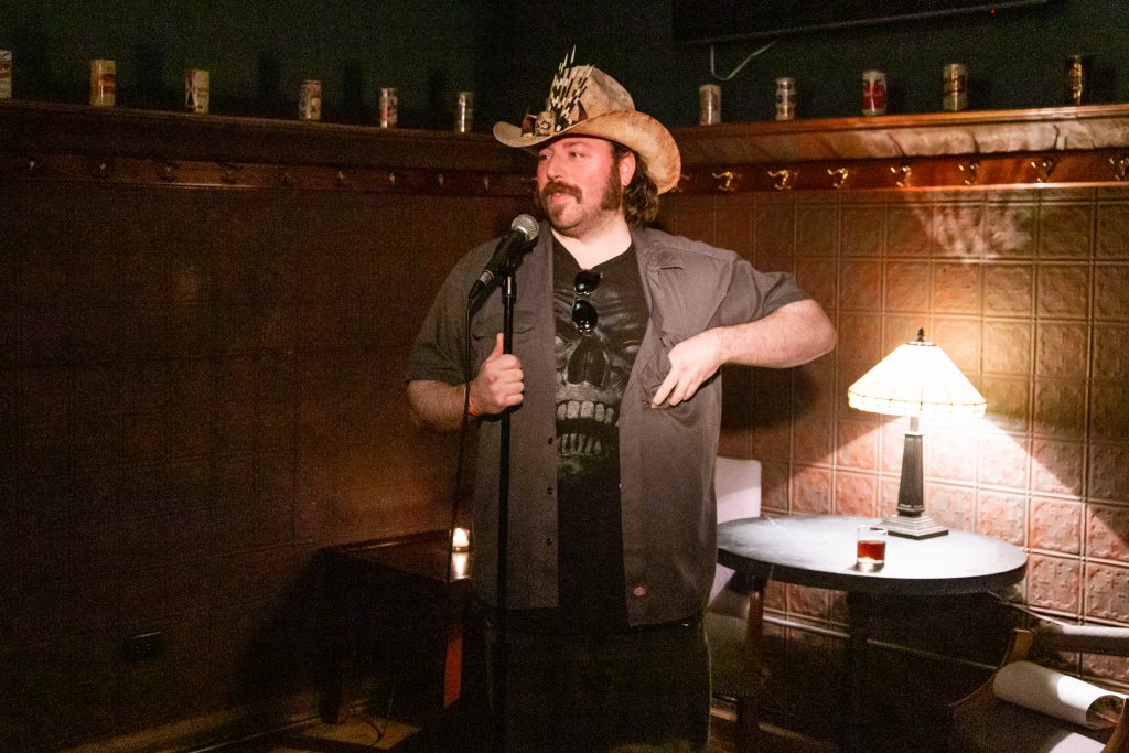 Image: J. Rohr performing at Unreal at Schubas. In this oblique, medium-long shot, Rohr stands at the front of the room, looking out at the audience. One of Rohr’s hands is holding the mic stand and the other is in Rohr’s shirt chest pocket. Behind Rohr is a copper-colored wall, made of a grid of low-relief tiles; above that are several decorative beer cans on a ledge and a dark green section of wall. Rohr wears a wide-brimmed hat with curved sides and small black, white, and brown objects attached at its front, as well as a grey short-sleeved collared shirt open over a black shirt that depicts a giant skull. Photo by Joshua Clay Johnson.
