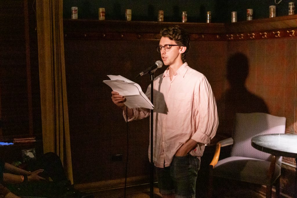 Image: Nick Russell performing at Unreal at Schubas. In this oblique, medium-length shot, Russell stands at the front of the room, speaking into a microphone while reading off a creased paper. Behind Russell is a copper-colored wall, made of a grid of low-relief tiles; above that are several decorative beer cans on a ledge and a dark green section of wall. Russell wears a pale pink collared shirt with sleeves rolled up, as well as dark-rimmed glasses. Photo by Joshua Clay Johnson.