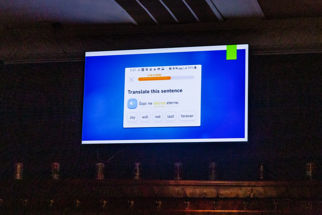 Image: An excerpt of a piece by Jerliyah Craig (not present), comprised of presentation slides being advanced by Chelsea Fiddyment (out of frame) at Unreal at Schubas. Against a blue background, this slide shows a phone screenshot of a translation activity from the app Duolingo, with the same sentence in Esperanto (“Ĝojo ne daŭros eterne”) and English (“Joy will not last forever”). The slides are being presented on a flat-screen monitor, above and below which parts of the wall and ceiling are visible. The room’s lights are off. Photo by Joshua Clay Johnson.