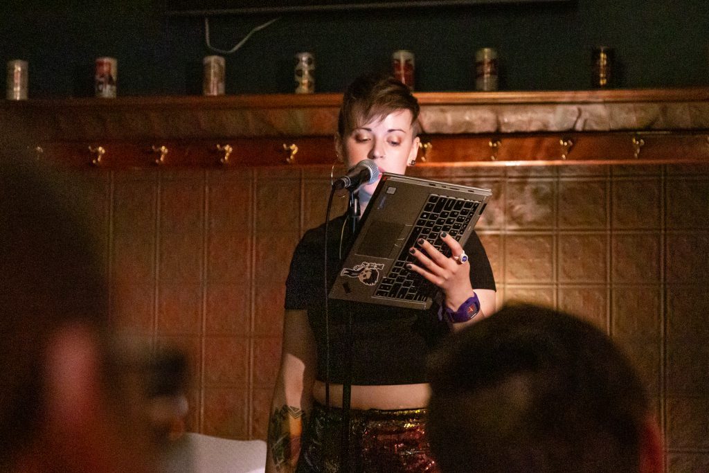 Image: Chelsea Fiddyment performing at Unreal at Schubas. Fiddyment stands at the front of the room, speaking into a microphone, with laptop in hand and folded over so the screen faces Fiddyment and the keyboard faces the audience. Behind Fiddyment is a copper-colored wall, made of a grid of low-relief tiles; above that are several decorative beer cans on a ledge and a dark green section of wall. Fiddyment wears a black cropped tee and red and gold sequined shorts. The backs of some audience members’ heads are fuzzy in the foreground. Photo by Joshua Clay Johnson.