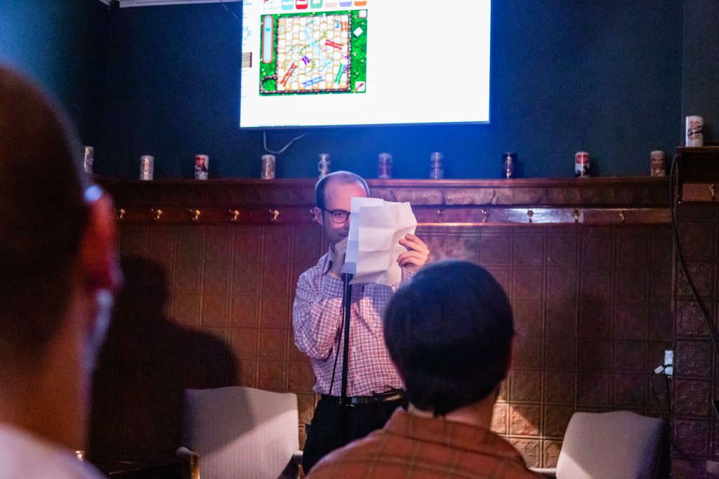 Image: John Pinkus performing at Unreal at Schubas. Pinkus stands at the microphone at the front of the room, face partially blocked by a creased paper. Above and behind Pinkus, on the wall, is a flat-screen monitor showing an internet-based game of Snakes and Ladders (being played for Pinkus by Chelsea Fiddyment, out of the frame). Also behind Pinkus is a copper-colored wall, made of a grid of low-relief tiles, and a door; above that are several decorative beer cans on a ledge and a dark green section of wall. Pinkus wears a white, blue, and orange collared shirt and dark-rimmed glasses. The back of some audience members’ heads are unfocused in the foreground. Photo by Joshua Clay Johnson.