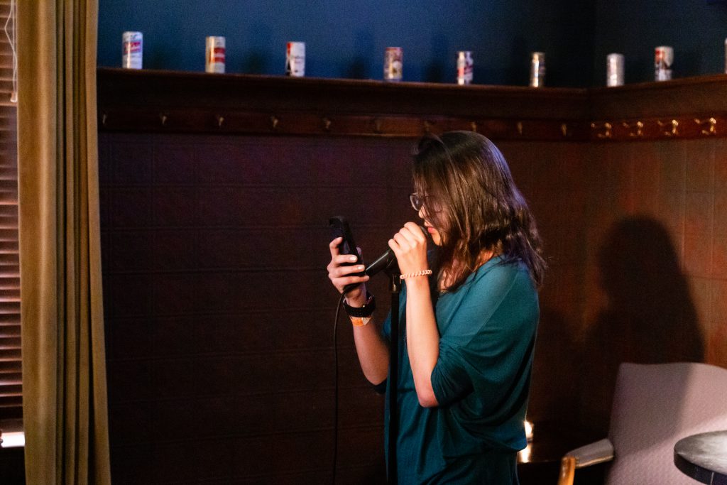 Image: Antonina Neva performing at Unreal at Schubas. In this oblique, medium-length shot, Neva stands at the front of the room, speaking into a microphone while reading off a phone. Behind Neva is a copper-colored wall, made of a grid of low-relief tiles; above that are several decorative beer cans on a ledge and a dark green section of wall. Neva wears a teal three-quarter sleeve shirt and dark-rimmed glasses. Photo by Joshua Clay Johnson.