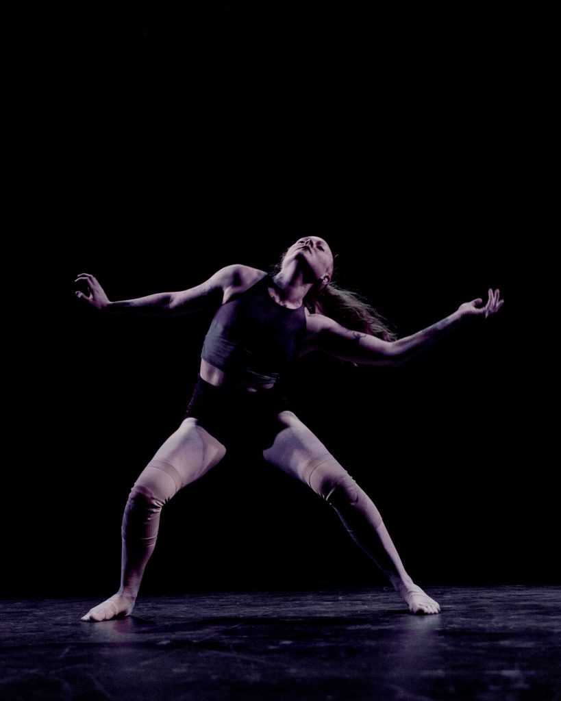  Image: A dancer (Julia Cox) dressed in a fitted top, shorts, and knee-wraps is in motion on a dark stage; she leans back, gesturing widely with her arms and legs splayed. Photo by Ryan Edmund.