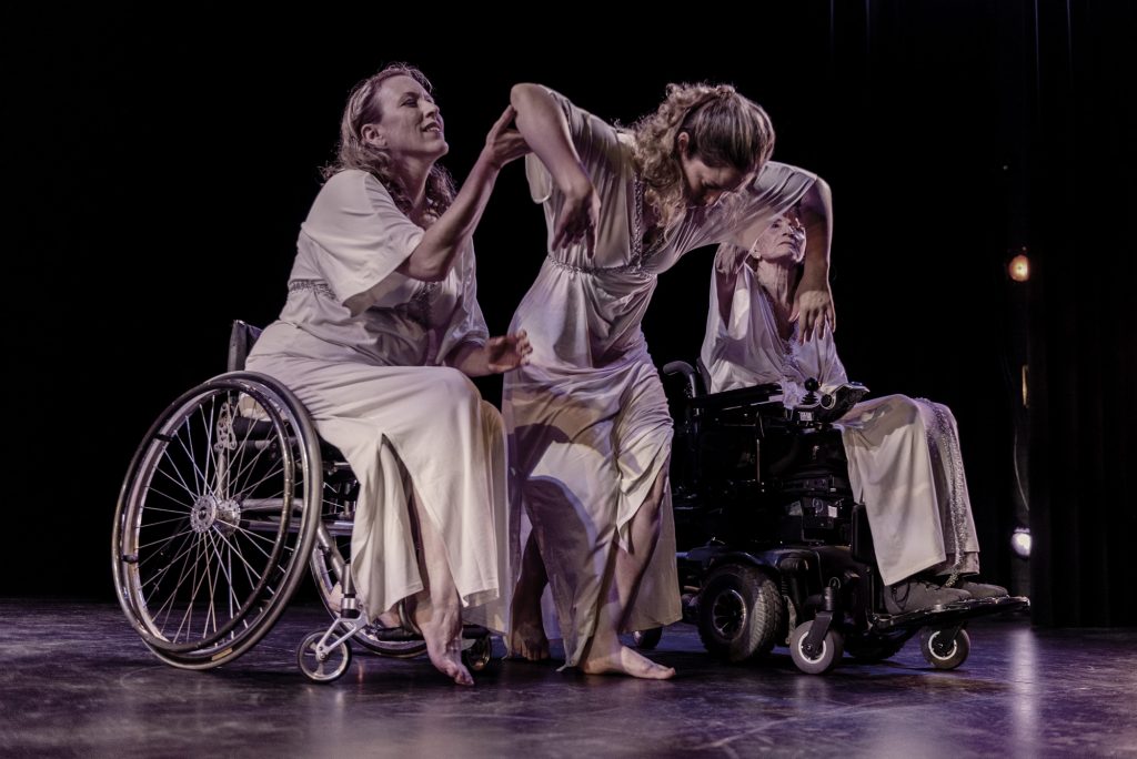 Image: Three dancers dressed in long white dresses are in motion on a dark stage; the dancer on the left (Ladonna Freidhem) is seated in an active wheelchair; the dancer on the right (Ginger Lane) is seated in a motorized wheelchair; the dancer at center (Anita Fillmore Kenney) has her head down, while her partners support her. Photo by Ryan Edmund.