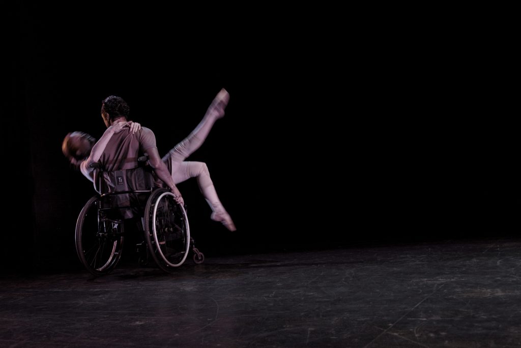 Image: Two dancers dressed in light grey clothing are in motion on a dark stage; one dancer (Julia Cox) reclines in the lap of the other dancer (Robby Williams), with her legs pointed expressively into the air; her partner is seated in an active wheelchair with his back to the camera. Photo by Ryan Edmund. 