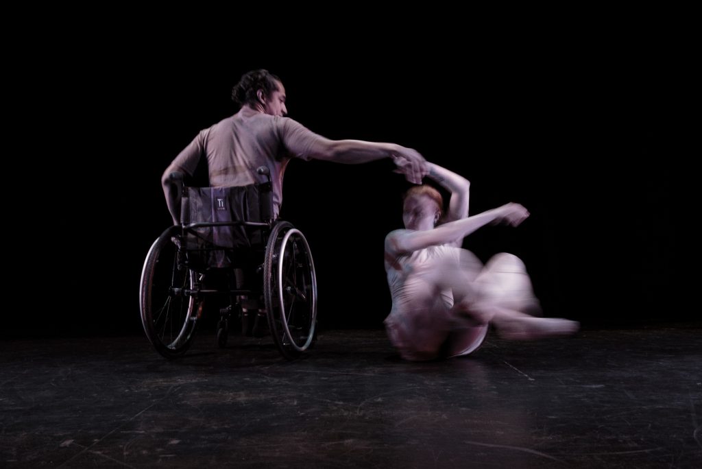 Image:  Two dancers dressed in light grey clothing are in motion on a dark stage; the dancer on the left (Robby Williams) is seated in an active wheelchair with his back to the camera; the dancer on the right (Julia Cox) spins on the floor, with the help of her partner. Photo by Ryan Edmund.