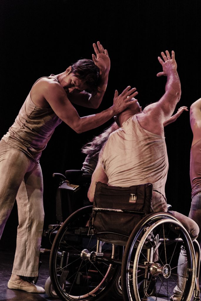  Image: Dancers dressed in light grey clothing are in motion on a dark stage; at left a dancer bows his head and lifts his hands, one hand is pressed against the back of another dancer's head; that dancer, seated in an active wheelchair, also raises his arm in a similar motion; other dancers can be seen in the background. Photo by Ryan Edmund.