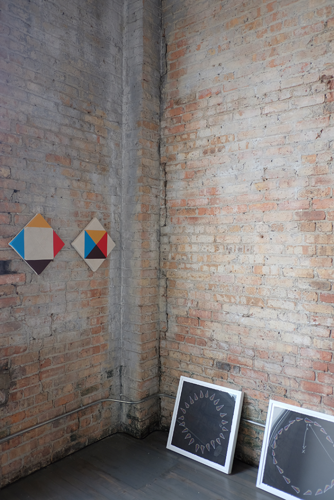 Image: The corners of a fading brick room meeting. On the left, about five feet off the ground are two canvases with an inch between them. These are Paolo Arao's "Greater Than (Diptych)" Leaning against the brick wall to the right on the floor are two of Jeffery Augustine Songco's "Choreography Playbook" series. Image courtesy of FLXST Contemporary.
