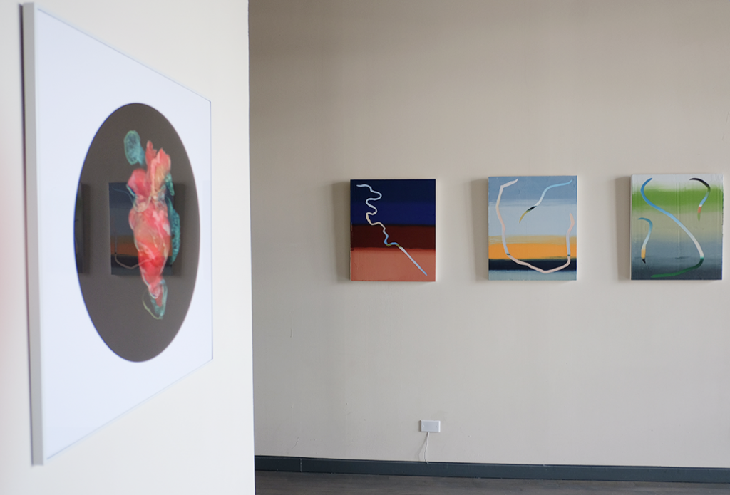 Image: A gallery with beige walls. Left, a square frame containing Kelvin Burzon's LATEX: Heart: a white drop containing a black circle containing a photograph of dyed latex and condoms approximating the shape of a heart. Right, on another wall and hung five feet off the ground,  Roberto Jamora's trio, from left to right, Bayou St. Malo, Lake Borgne, and Manila Plaza. Image courtesy of FLXST Contemporary.
