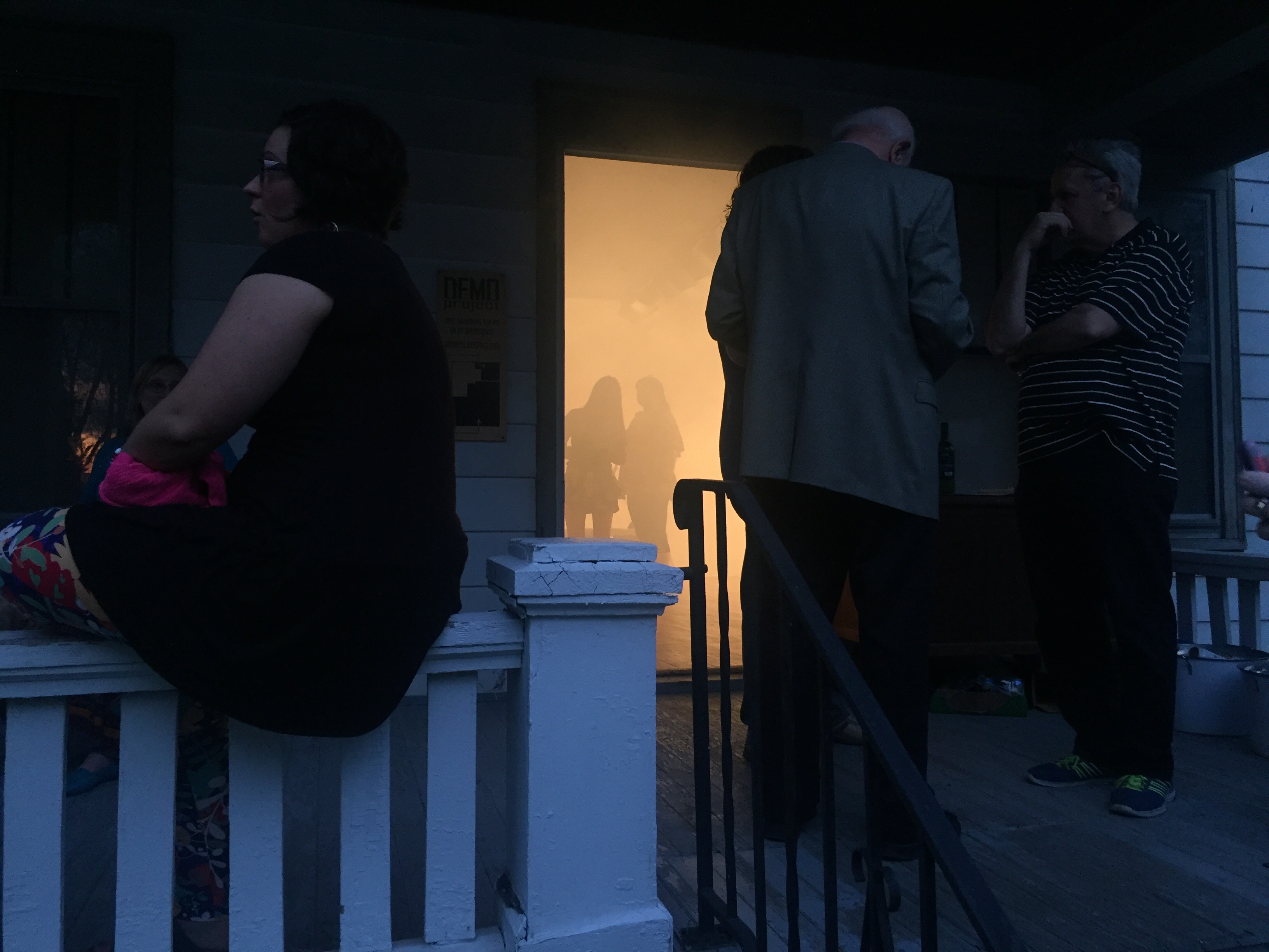 Image: Reception and entrance to Nobody's Home by Lyndon Barrois Jr., Addoley Dzegede, Cole Lu, and Catalina Ouyang. DEMO Project, May 2017. In the foreground, a few visitors linger on the front porch at dusk. Through the open doorway in the background, two figures stand viewing the exhibition through the orange haze. The Image courtesy of Lyndon Barrois Jr.