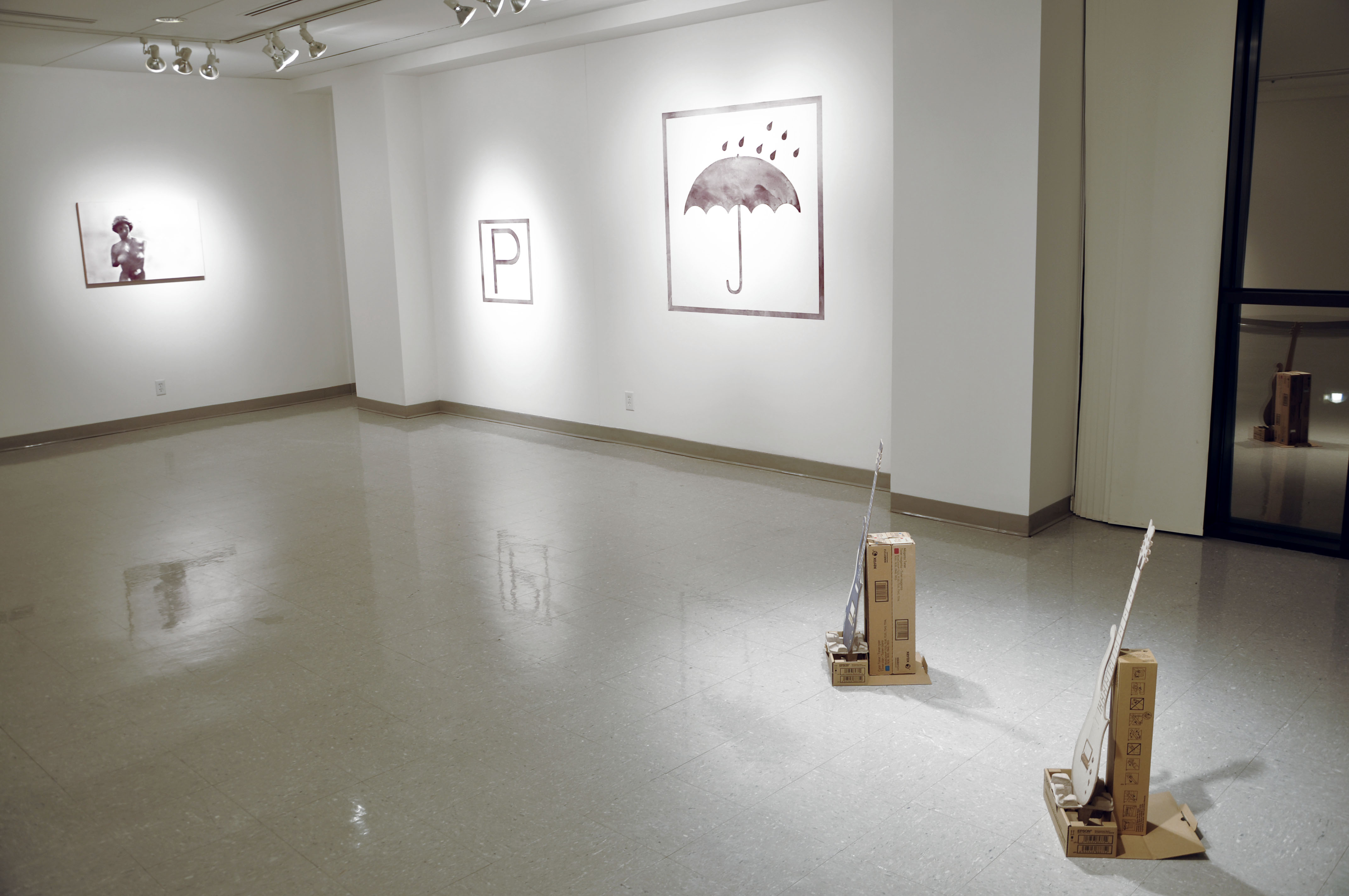 Image: Avez Vous un Crayon? (installation view) by Lyndon Barrois Jr. University of Illinois Springfield Visual Arts Gallery, September 2016. Two guitar cutouts lean against cardboard-like boxes on the gallery floor and three pieces hang on the gallery walls; one features an umbrella with raindrops, a smaller one of the letter ‘P’ and the third of the silhouette of a figure. Image courtesy of Lyndon Barrois Jr.
