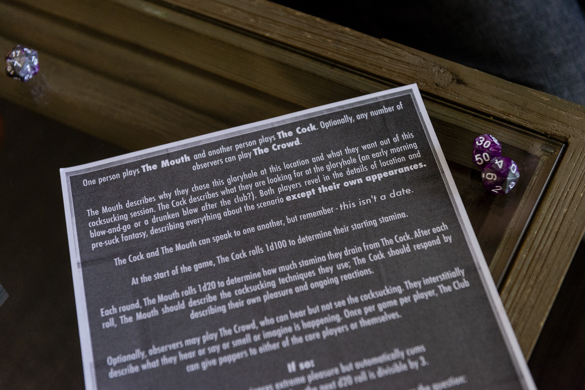 Image: A close up of a piece of paper with a black background and white text explains the game, "Welcome to Paradise, Baby" where one player is The Mouth, the other player is The Cock and observers, like our staff photographer, was The Crowd. To the right side of the paper are two die. Photo by Ryan Edmund Thiel